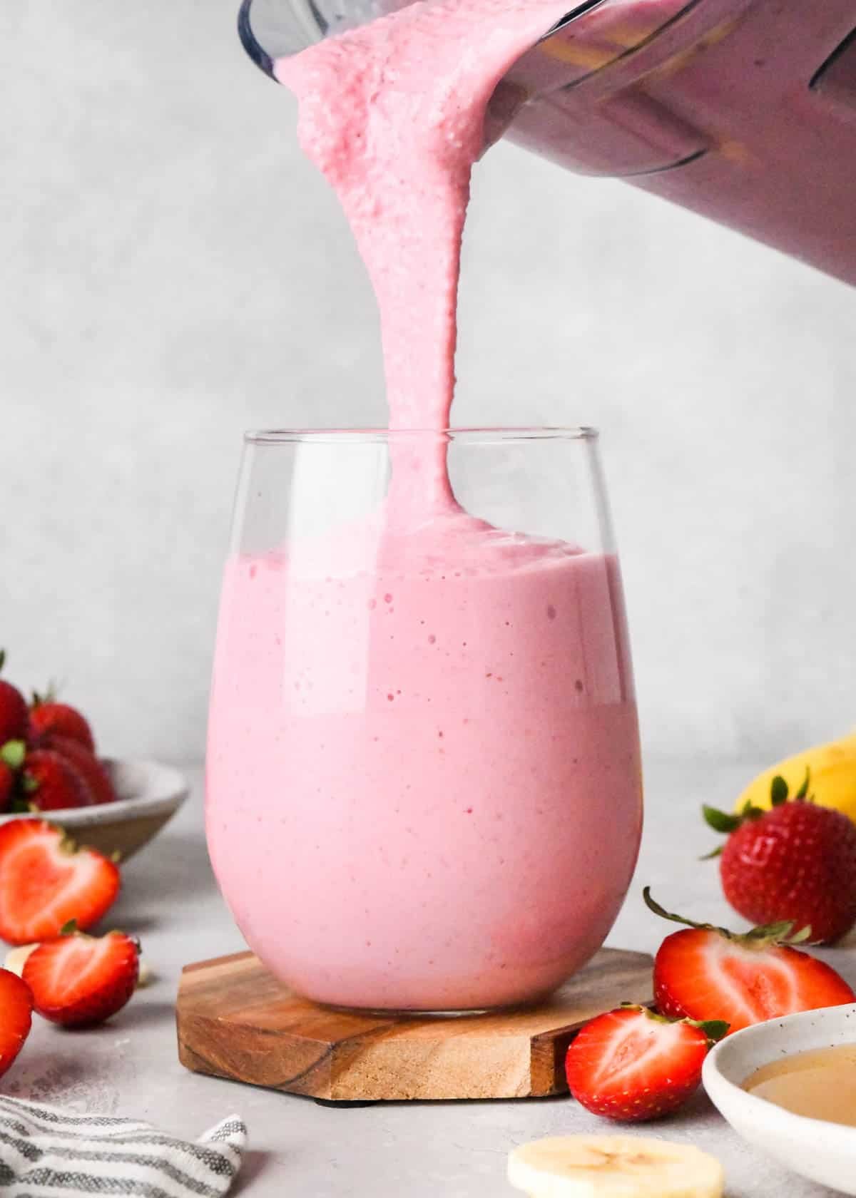 The BEST strawberry banana smoothie recipe ever! It's easy (ready in less than 5 minutes), healthy, and made with yogurt so it's full of protein! It's the perfect snack or breakfast! #smoothie #strawberrybananasmoothie #strawberry #banana #breakfast #snack #vitamix strawberry banana smoothie being poured from a blending container into a glass which is almost full of the smoothie. Surrounded by strawberries and bananas the glass is on a wooden coaster