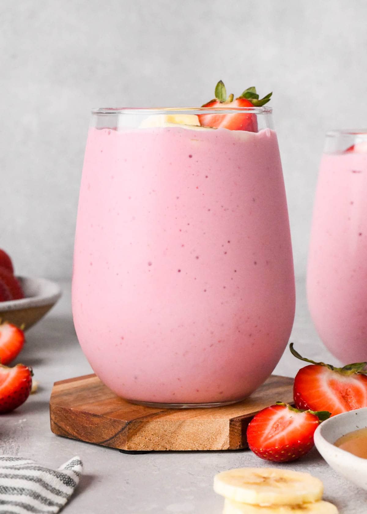 The BEST strawberry banana smoothie recipe ever! It's easy (ready in less than 5 minutes), healthy, and made with yogurt so it's full of protein! It's the perfect snack or breakfast! #smoothie #strawberrybananasmoothie #strawberry #banana #breakfast #snack #vitamix front view of strawberry smoothie in a glass topped with sliced bananas and sliced strawberries, there is another smoothie in a glass next to it, strawberries and bananas in the background