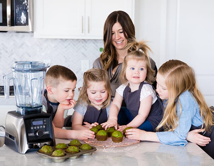 front view of a mom and her five kids reaching for muffins they made together while she was Front view of a mom and her four kids blending in a blender while she's Cooking with Kids