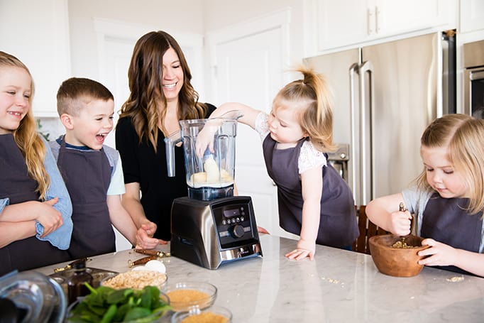 Front view of a mom and her four kids, the littlest one adding a banana to the blender while she's Cooking with Kids