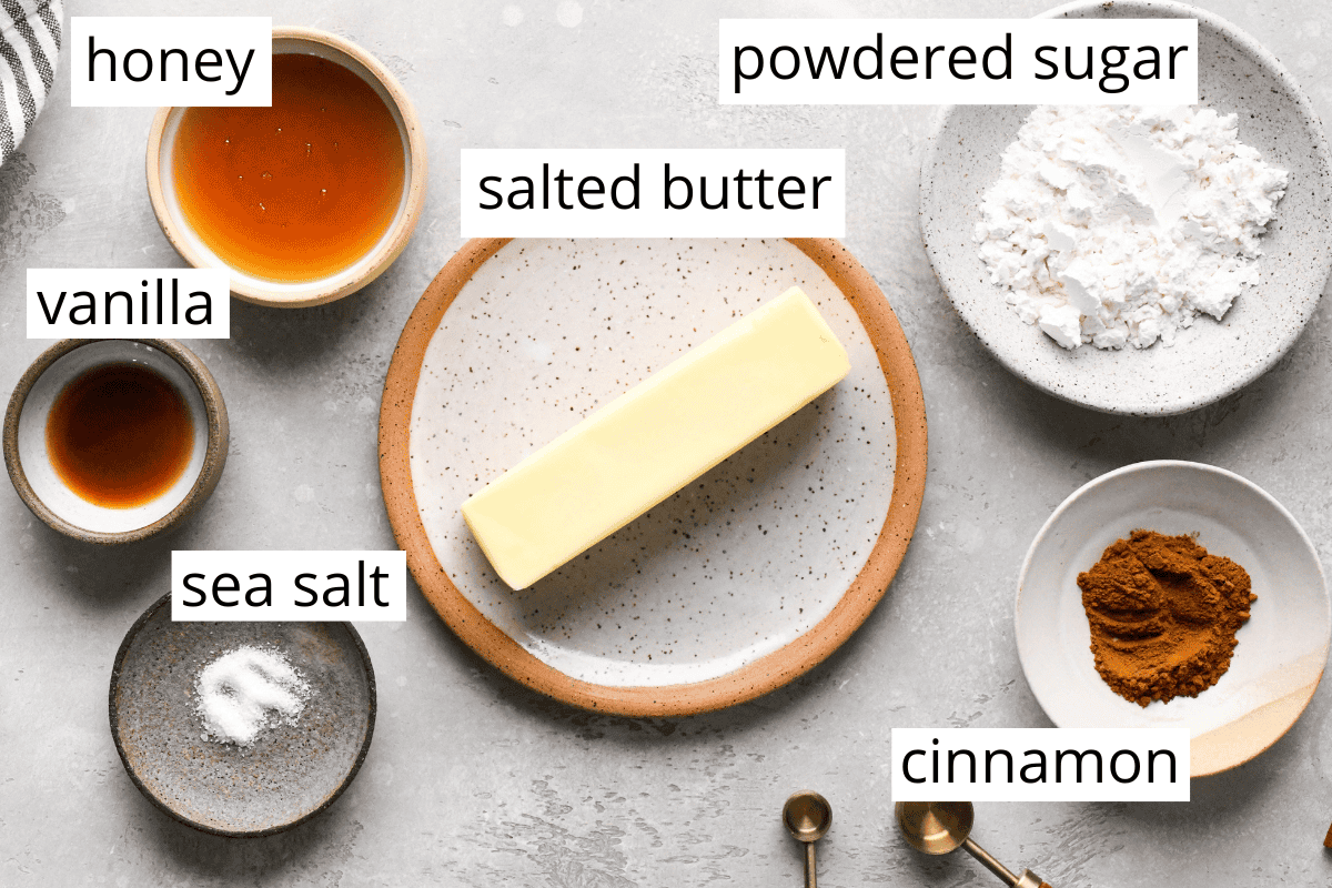 overhead photo of the ingredients in this cinnamon honey butter recipe in bowls and labeled, ingredients are honey, vanilla, powdered sugar, sea salt, cinnamon and butter.