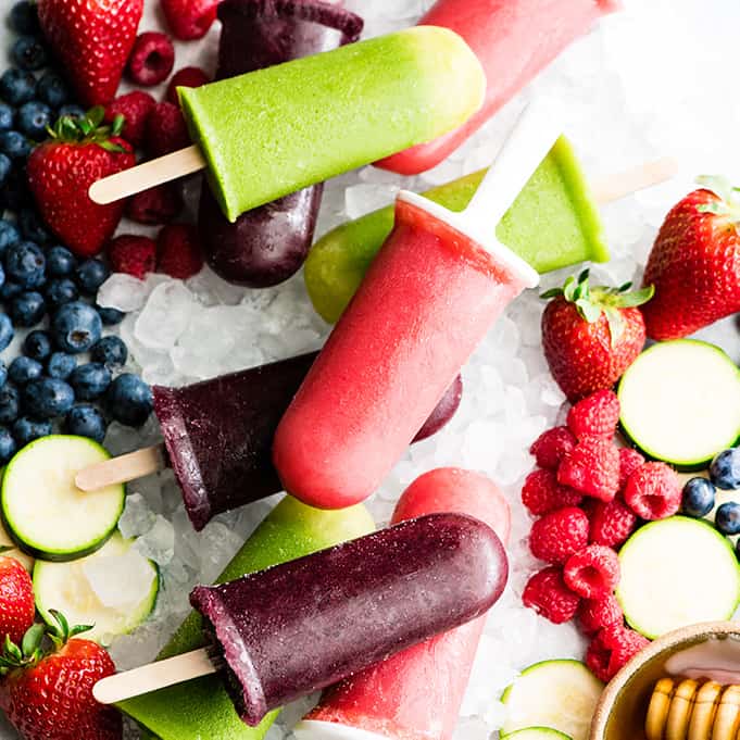 overhead view of 9 Homemade Fruit Popsicles in three colors: purple, green and red. Laying out on crushed ice with fresh fruit surrounding them.