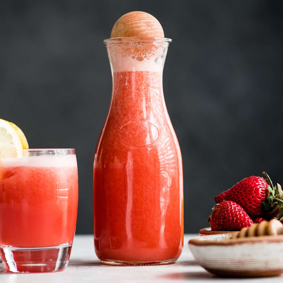 front view of a glass pitcher of Homemade Strawberry Lemonade and a glass of Homemade Strawberry Lemonade and a bowl of strawberries next to it 