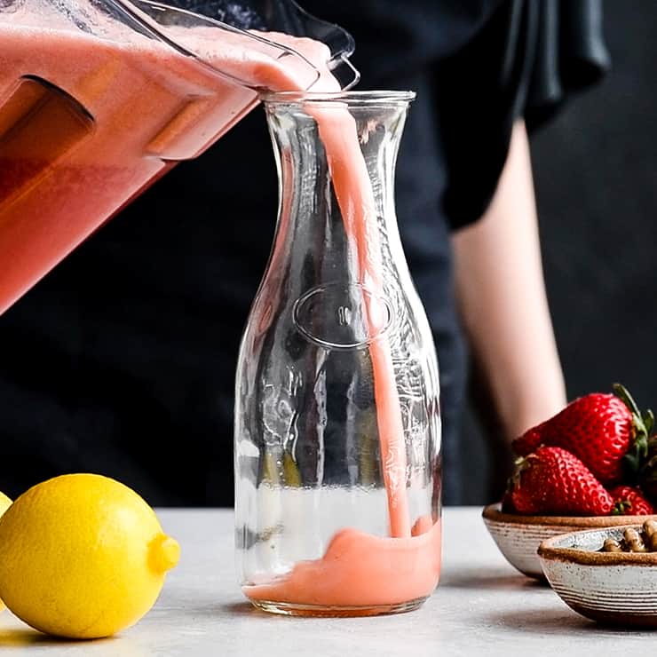 front view of a woman pouring Homemade Strawberry Lemonade from the vitamix container into a glass pitcher. 