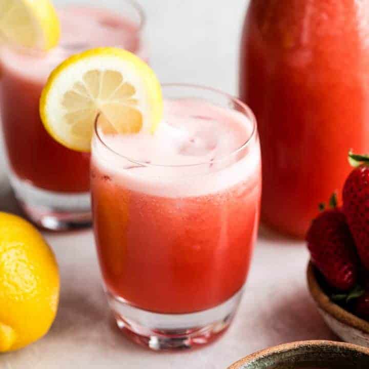 front view of a glass of Homemade Strawberry Lemonade with a lemon slice on the rim. Another glass and a pitcher of Homemade Strawberry Lemonade sit behind the front glass on the left and right. two lemons are sitting on the left side and a bowl of strawberries on the right. 