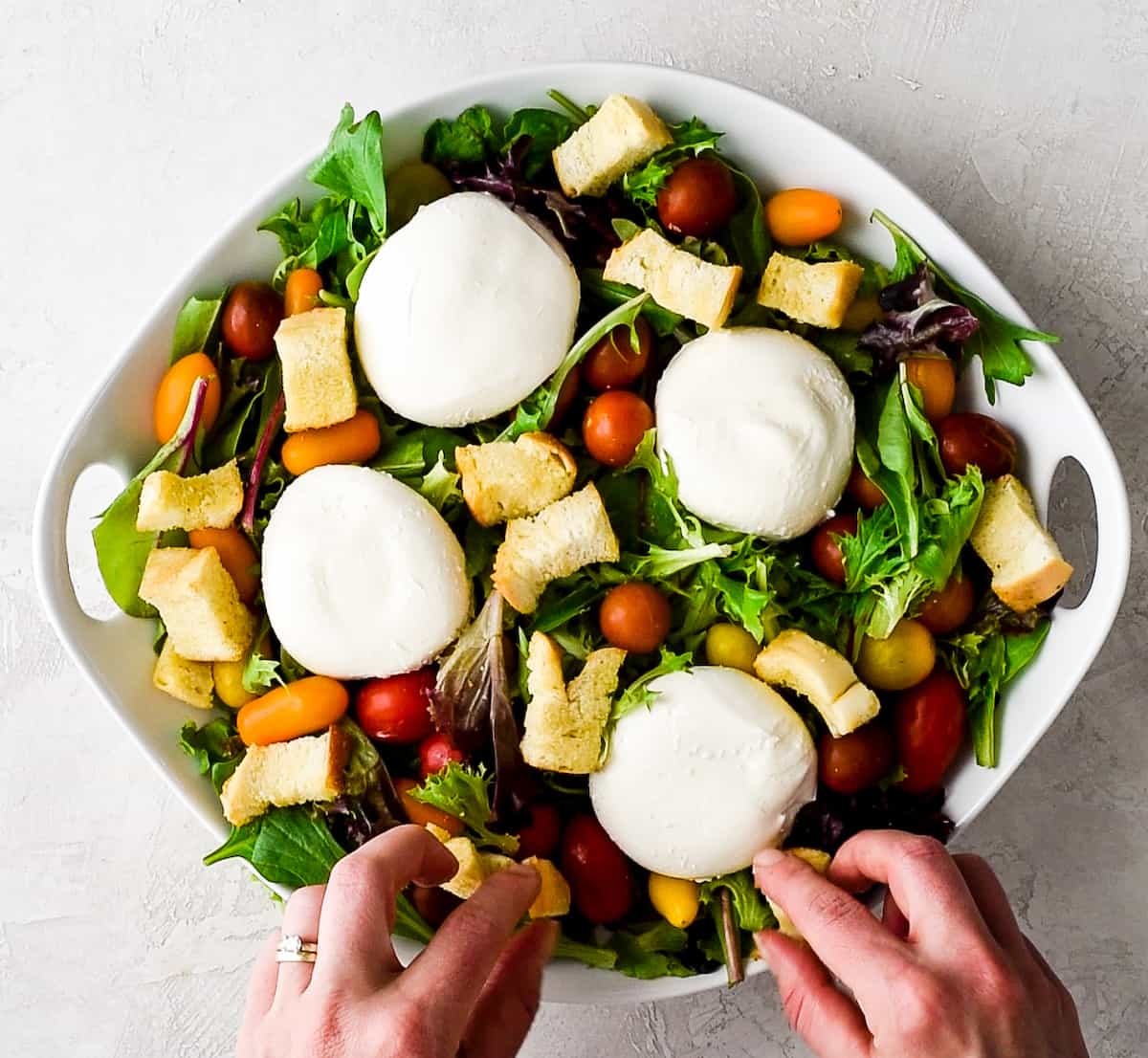 overhead view of a large white bowl with spring mix, baby tomatoes, four burrata cheese rounds and hands putting croutons on top of the greens making this burrata salad