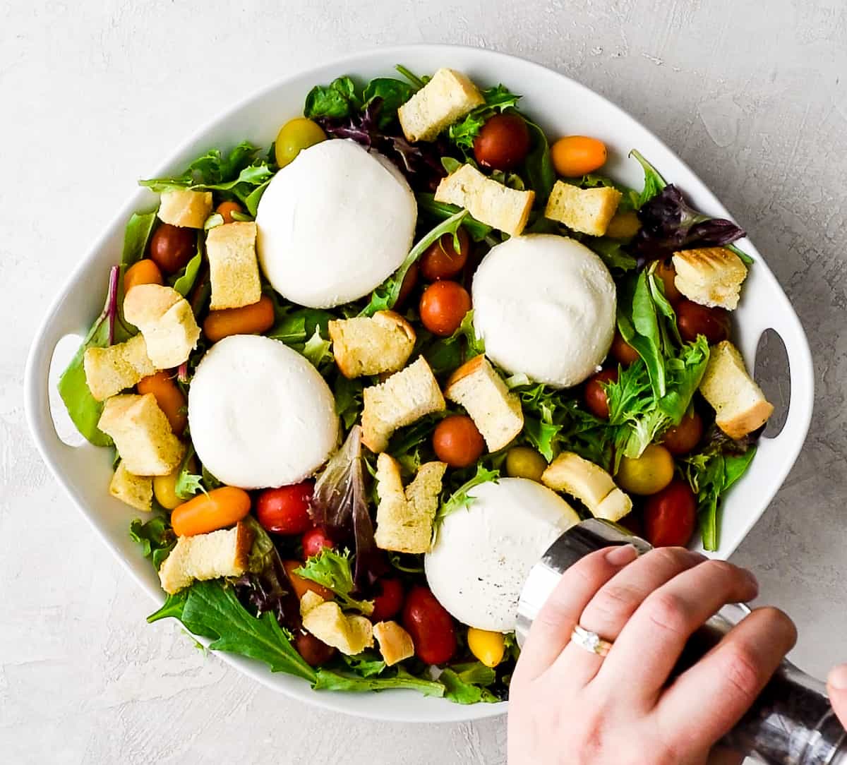 overhead view of hands grinding pepper on the top of a burrata cheese round that's on a large white bowl with spring mix, baby tomatoes, four burrata cheese rounds and homemade croutons - a burrata salad