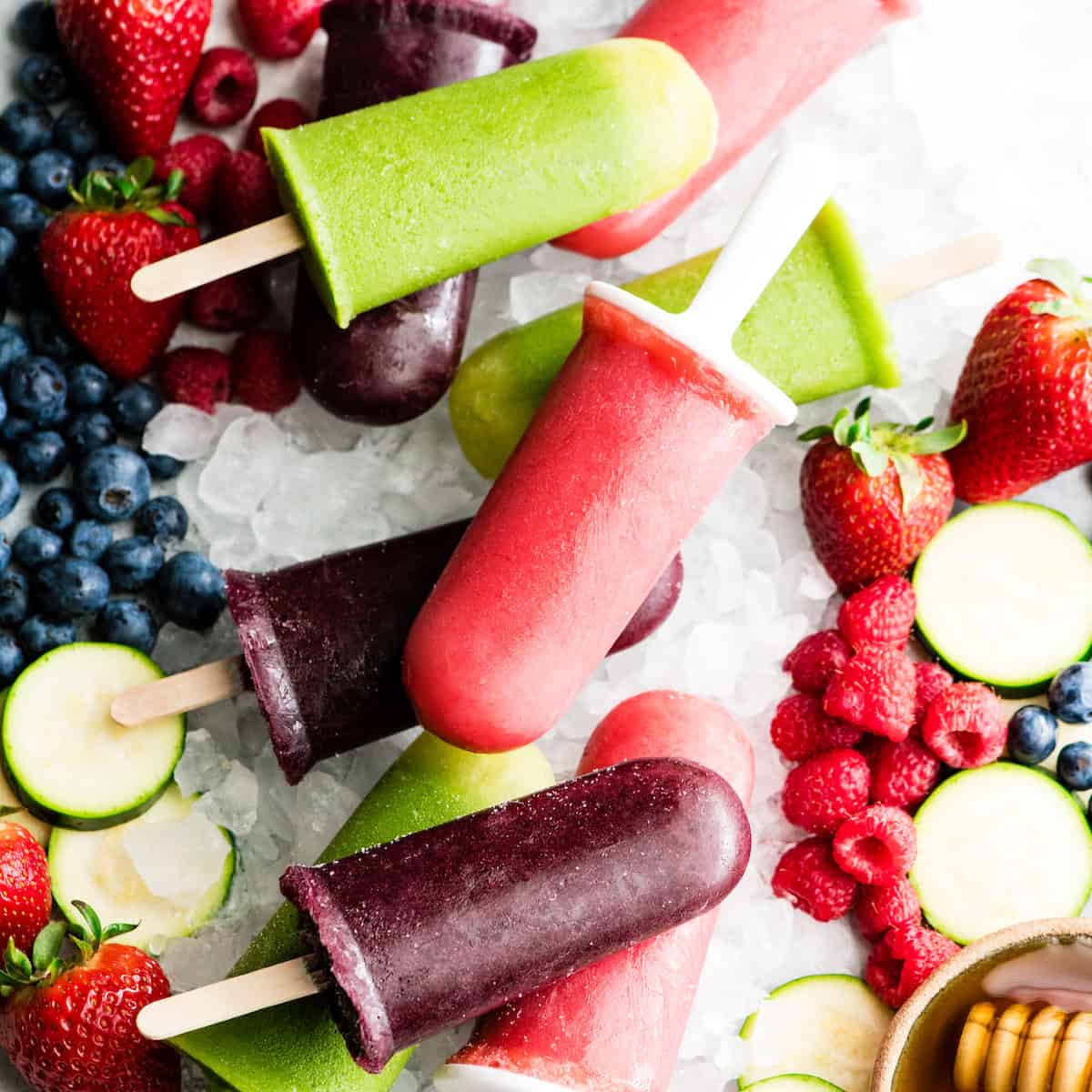overhead view of 9 Homemade Fruit Popsicles in three colors: purple, green and red. Laying out on crushed ice with fresh fruit surrounding them.