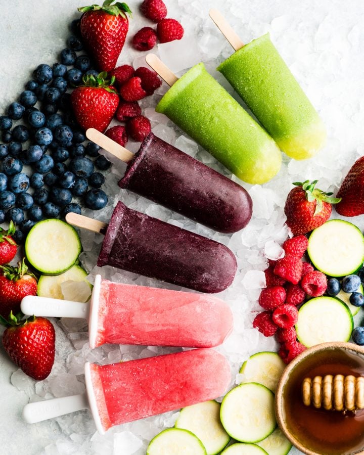 overhead view of 6 Homemade Fruit Popsicles in three colors: purple, green and red. Laying out on crushed ice with fresh fruit surrounding them.