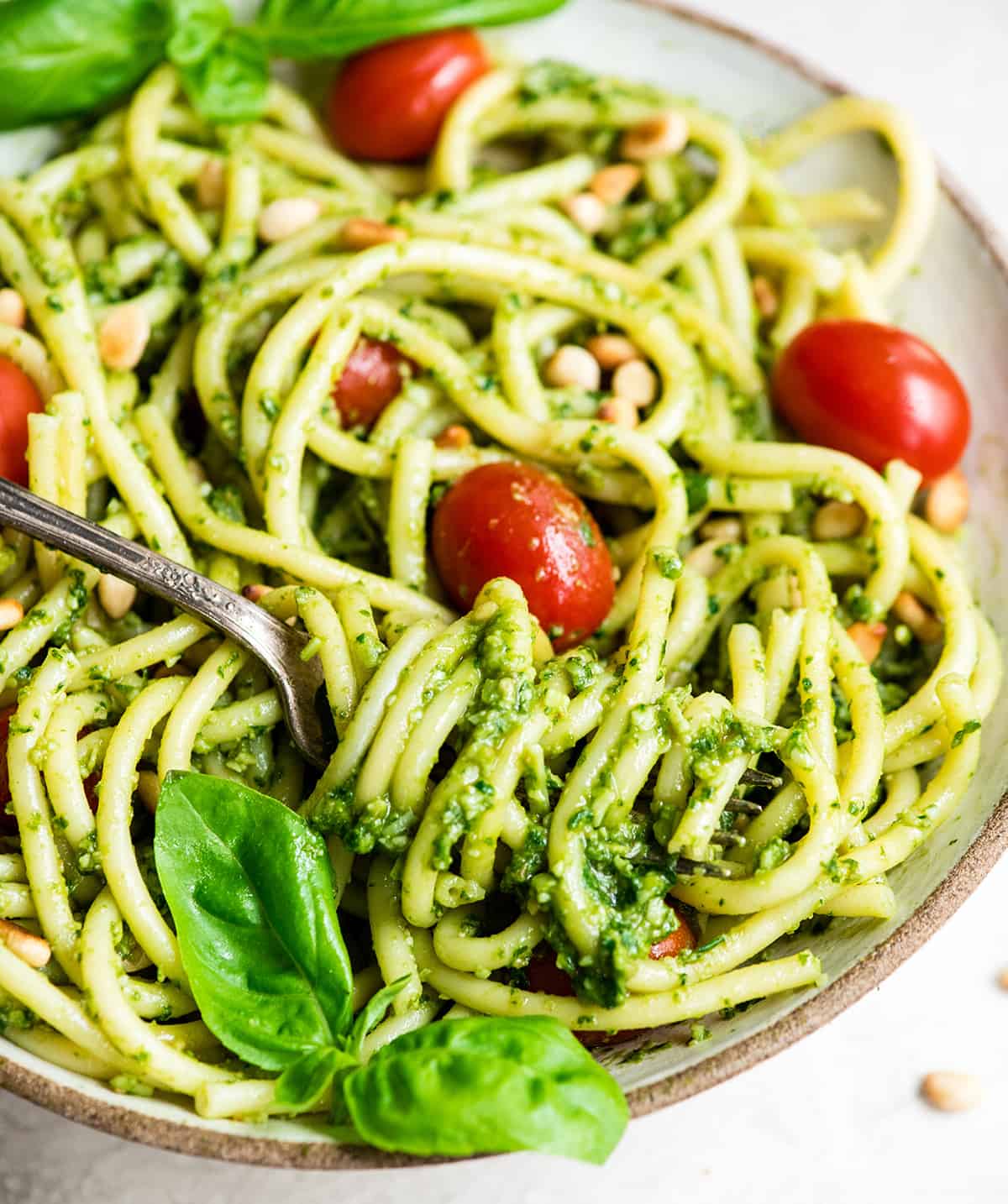 up close front view of a bowl of pasta with Basil Pesto Sauce on it garnished with fresh basil leaves, toasted pine nuts, and red cherry tomatoes. There is a fork with pasta twirled on it. 