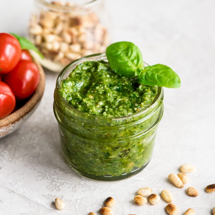 front overhead view of a small glass jar of Basil Pesto Sauce with two fresh basil leaves on the top surrounded by toasted pine nuts and a bowl of cherry tomatoes