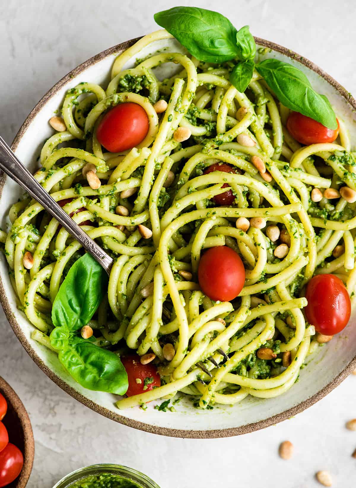overhead view of a bowl of pasta with Basil Pesto Sauce on it garnished with fresh basil leaves, toasted pine nuts, and red cherry tomatoes. There is a fork with pasta twirled on it. 