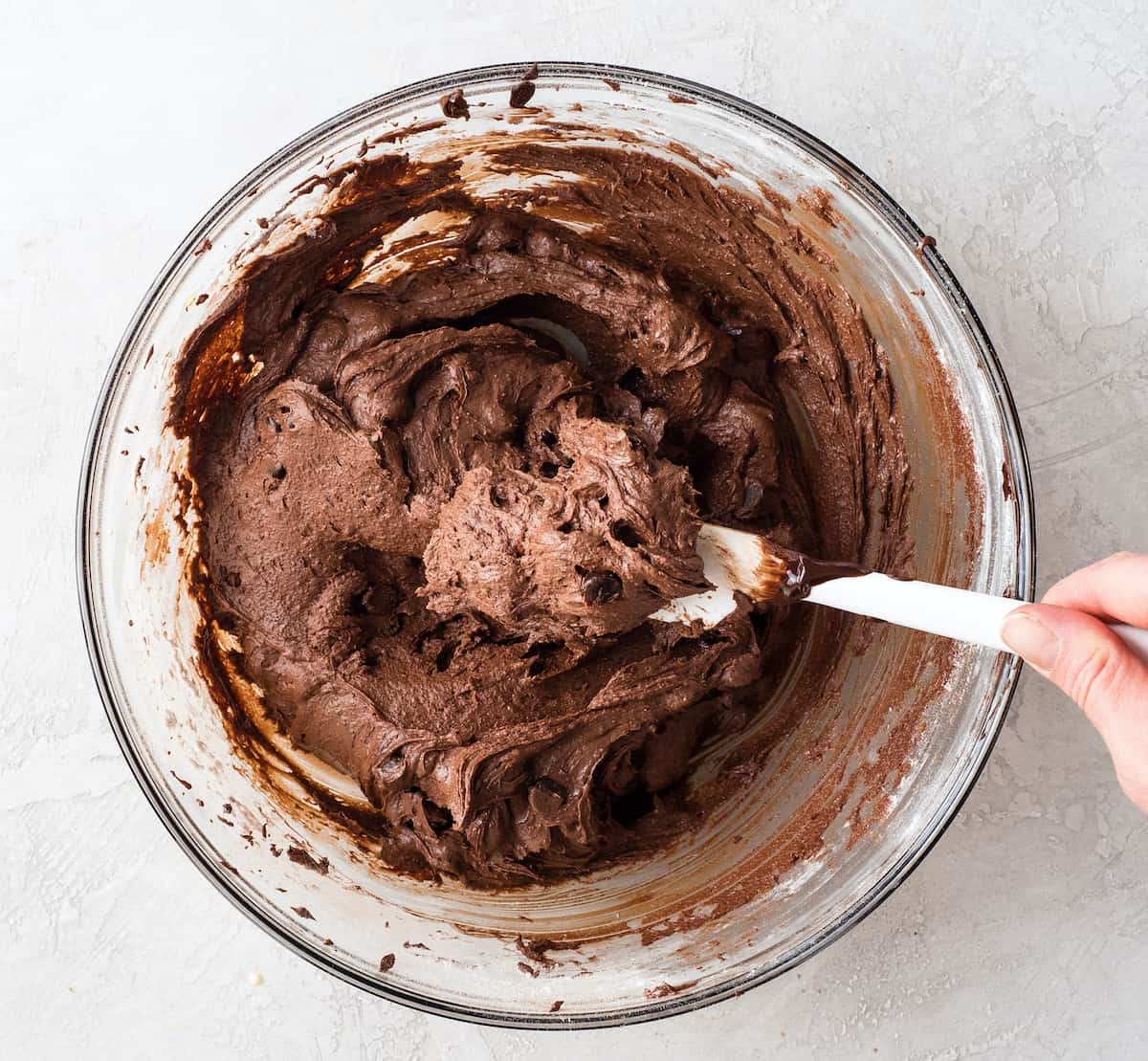 overhead photo of a hand holding a white spatula in a glass bowl of Brownie Recipe batter to show the thickness