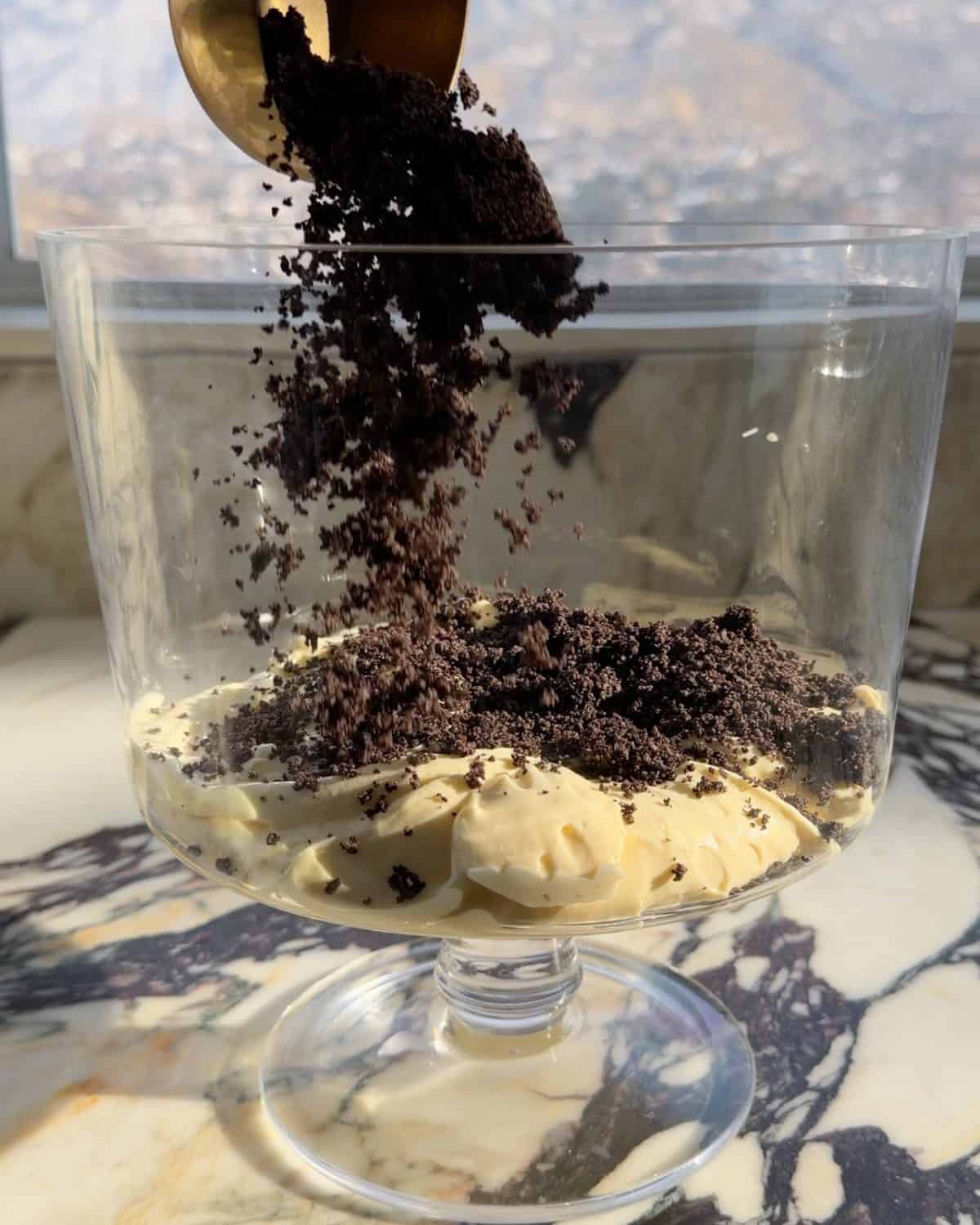 assembling this dirt cake recipe - the first layer of oreo crumbs being poured over the first layer of pudding in a glass trifle dish