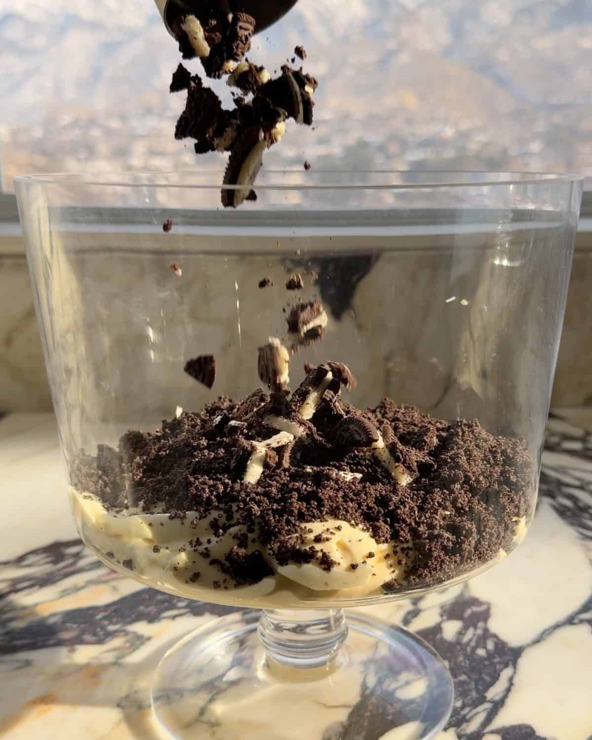 assembling this dirt cake recipe - first layer of chopped cookies being sprinkled over the first layer of cookie crumbs in a glass trifle dish on a marble surface