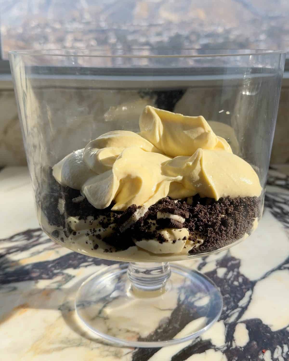 assembling this dirt cake recipe - the second layer of pudding being added on top of the chopped cookie layer in a glass trifle dish on a marble surface 