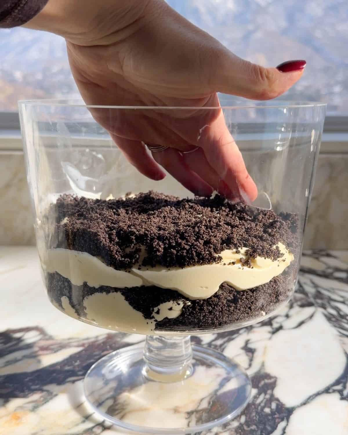 assembling this dirt cake recipe - a hand spreading out the second layer of cookie crumbs in a glass trifle dish on a marble surface