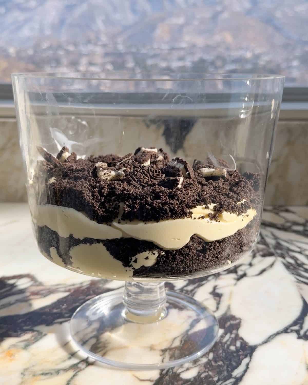 assembling this dirt cake recipe - second layer of chopped oreo cookies added on top of the second cookie crumb layer in a glass trifle dish on a marble surface