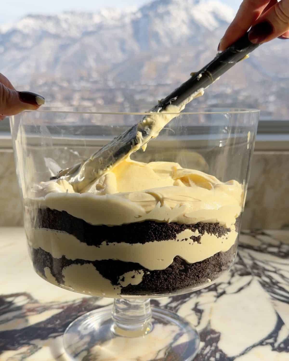 assembling this dirt cake recipe - a spatula spreading the third pudding layer into a glass trifle dish on a marble surface 