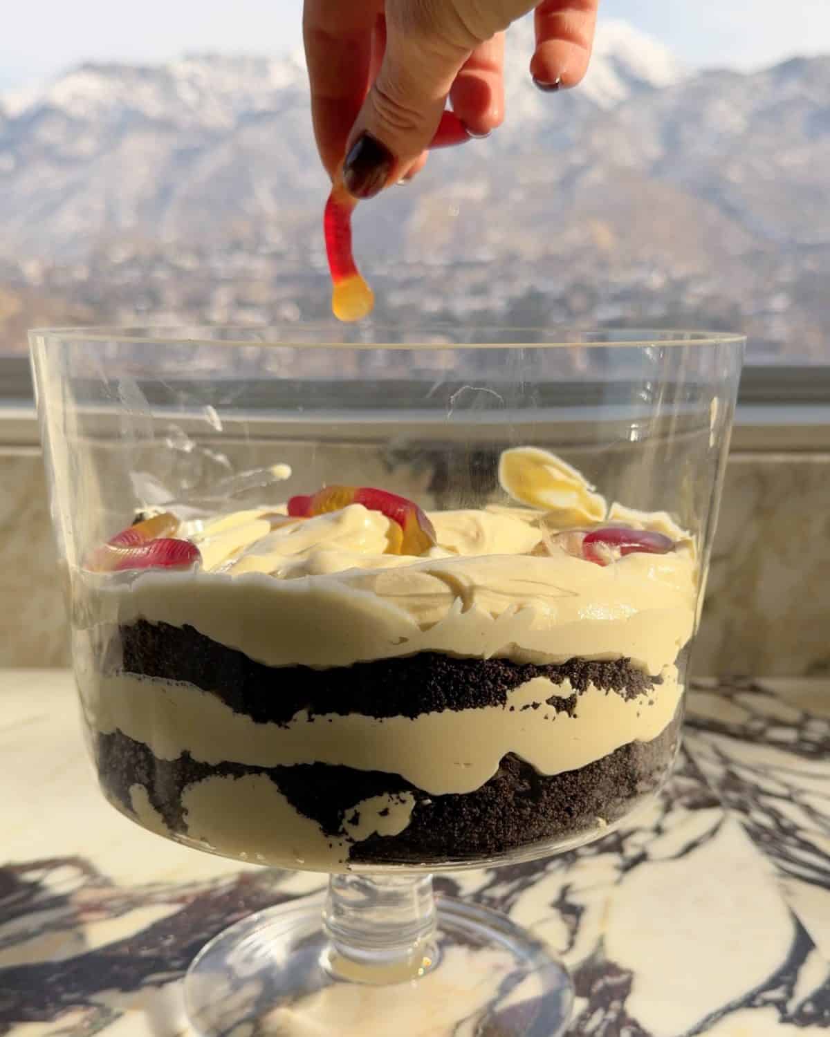 assembling this dirt cake recipe - a hand sprinkling gummy worms over the third pudding layer in a glass trifle dish on a marble surface