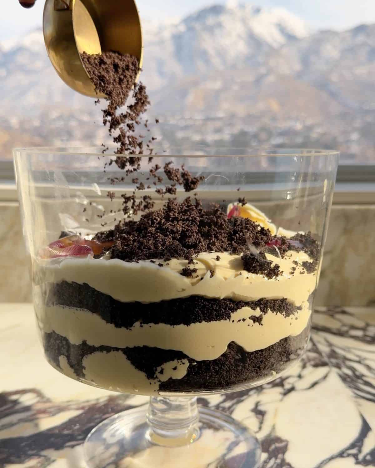 assembling this dirt cake recipe - the third layer of oreo cookie crumbs being poured over the gummy worm layer in a glass trifle dish on a marble surface