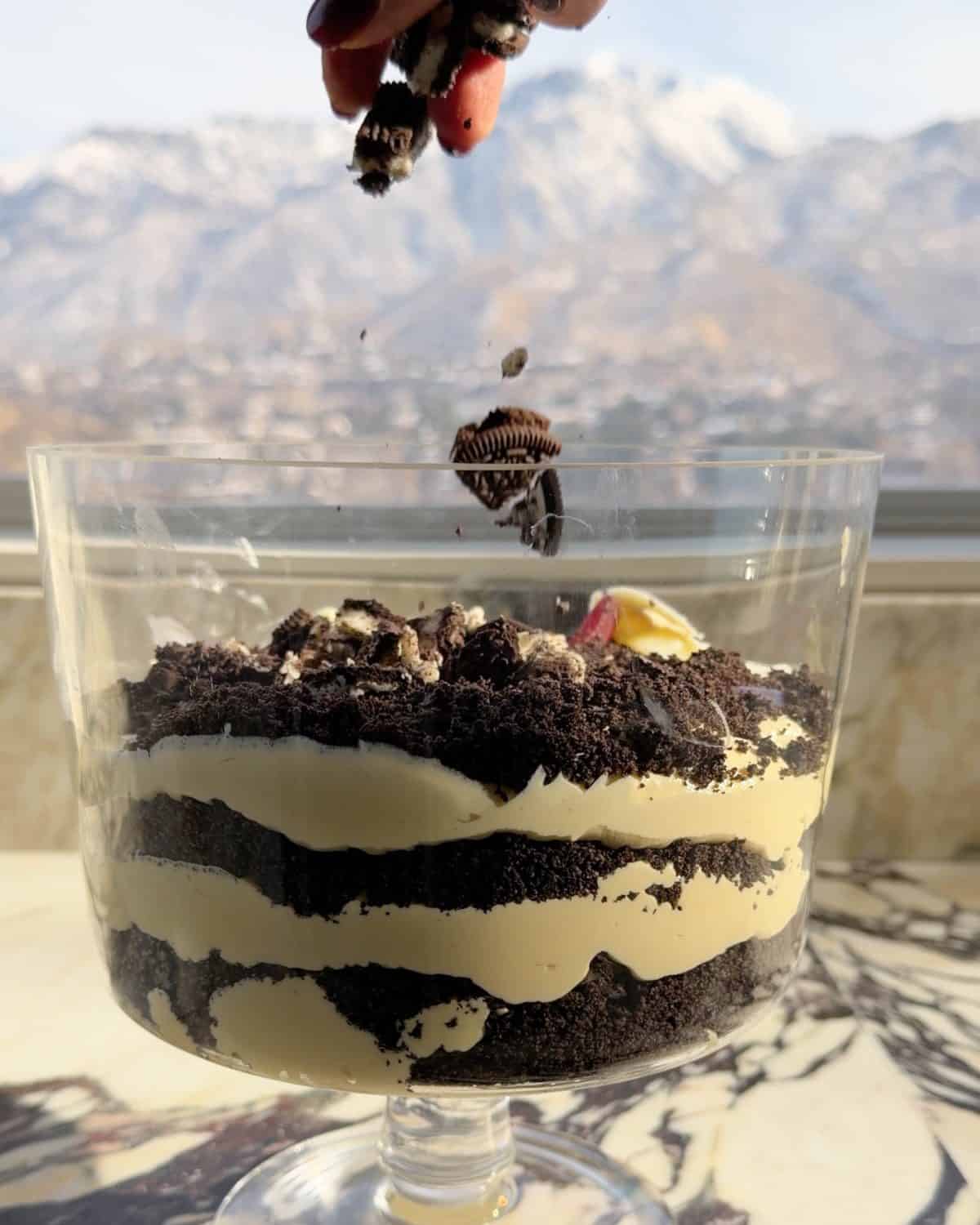 assembling this dirt cake recipe - a hand sprinkling the third layer of chopped oreo cookies into a glass trifle dish on a marble surface 