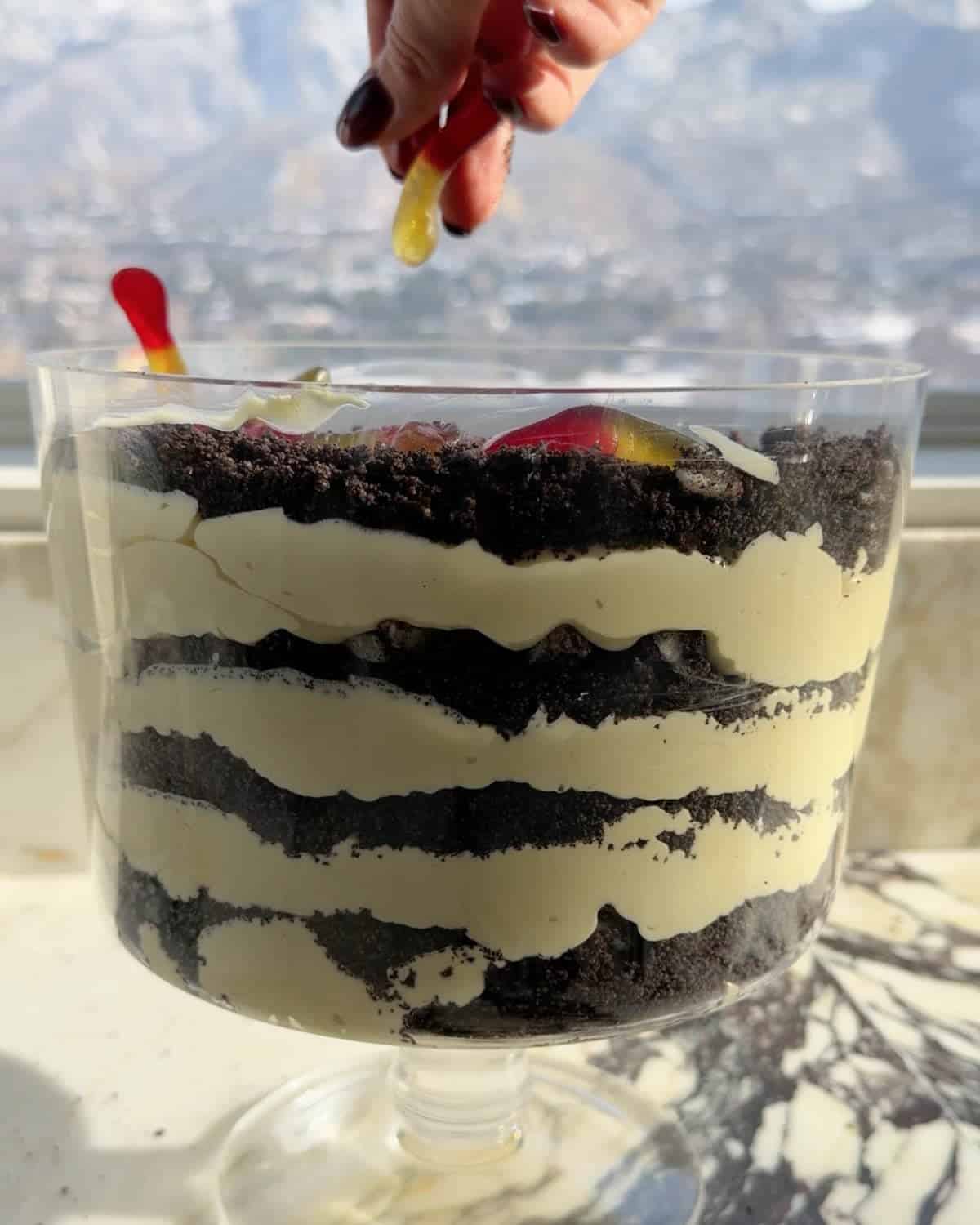 assembling this dirt cake recipe - final layer of gummy worms being added to the top of the dirt pudding in a glass trifle dish on a marble surface 
