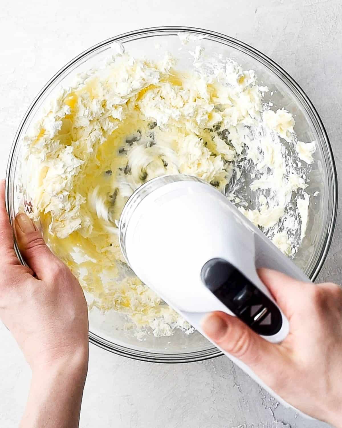 making oreo dirt cake recipe - hands beating butter and cream cheese together with a white handheld mixer in a large glass bowl on a gray surface