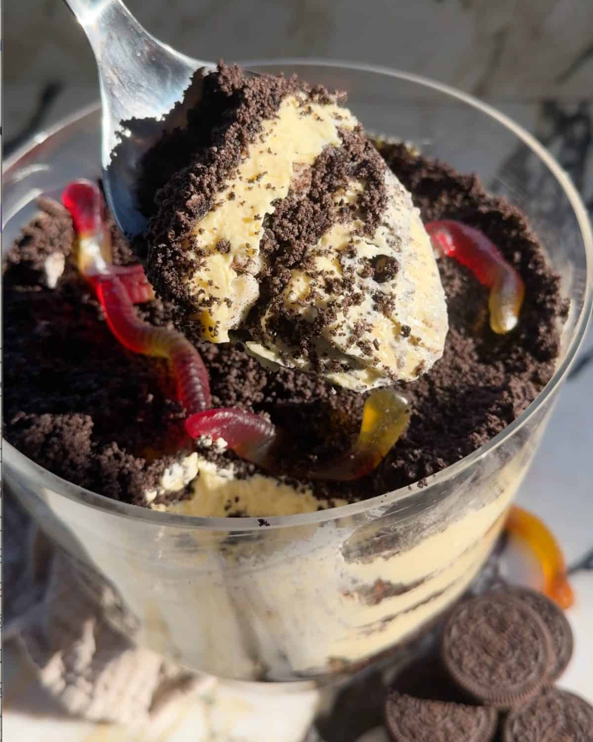 a metal spoon with a scoop of dirt cake on it showing 3 layers of pudding and cookies being held over the trifle dish with the rest of the dirt cake and gummy worms. 