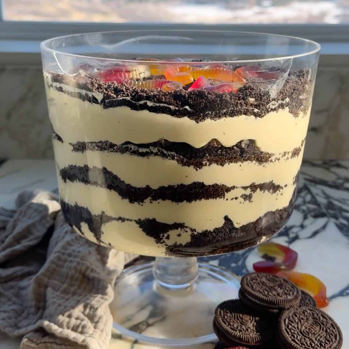Front view of this Oreo Dirt Cake Recipe in a glass trifle dish with 4 layers of pudding and 4 layers of crushed Oreos with gummy worms on top with oreos and gummy worms around the trifle dish