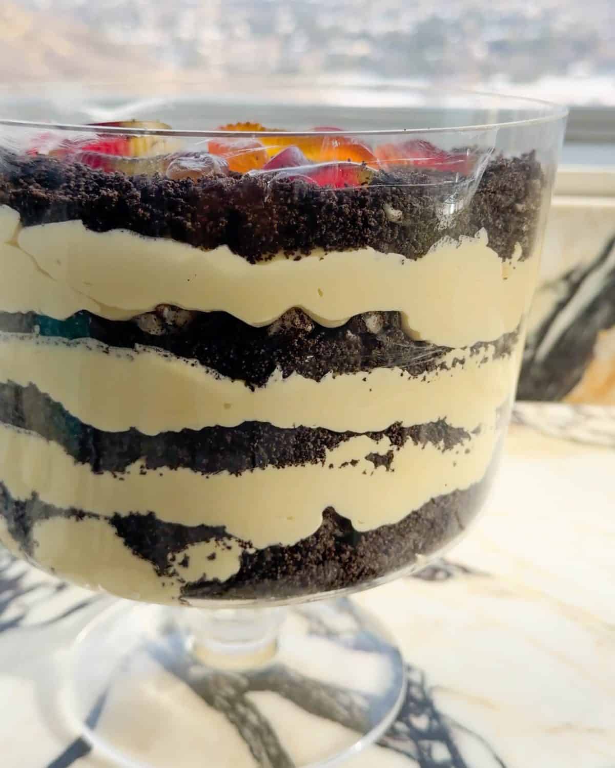 Front close up photo of this Oreo Dirt Cake Recipe in a glass trifle dish with 4 layers of pudding and 4 layers of crushed Oreos with gummy worms on top with oreos and gummy worms around the trifle dish