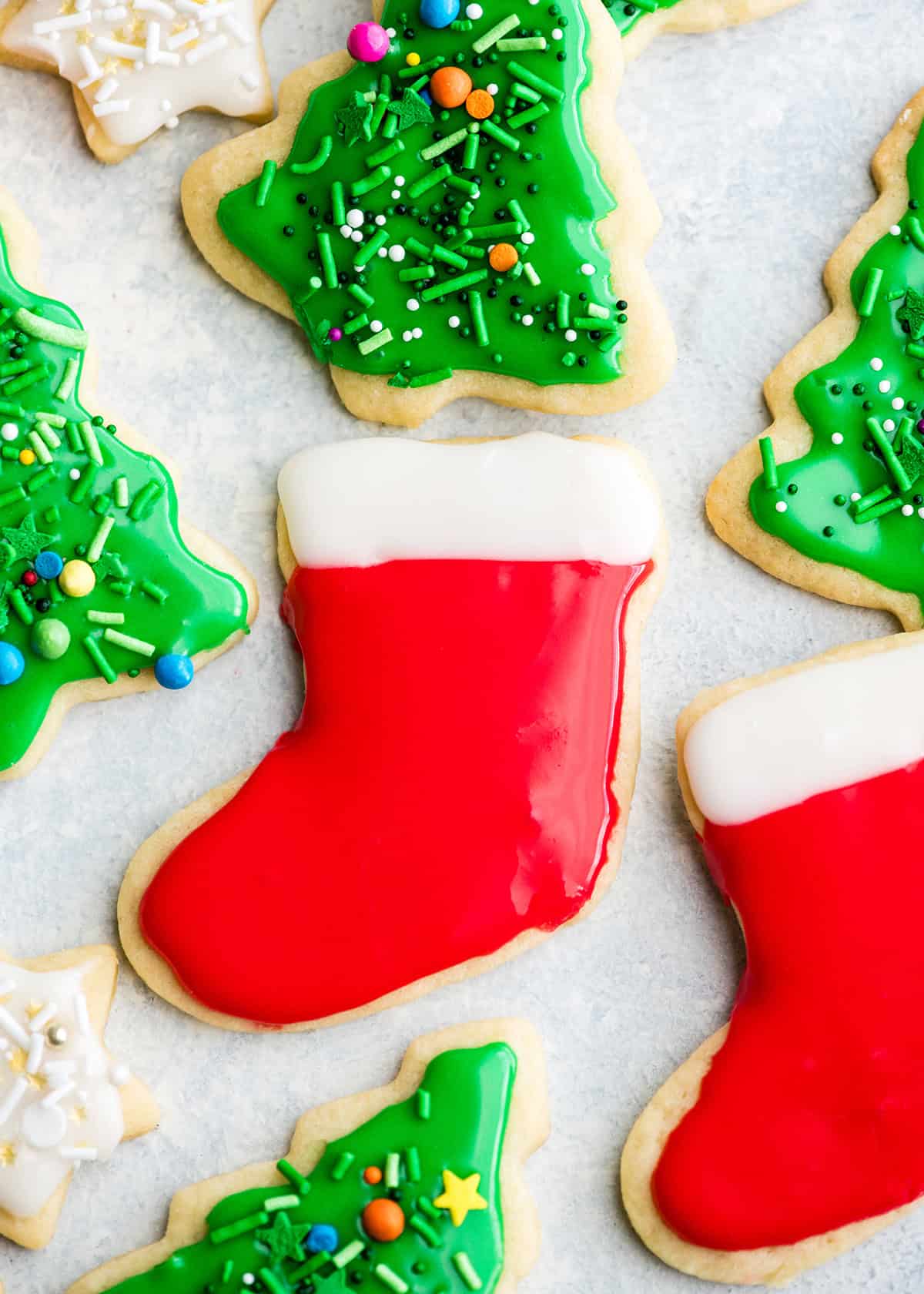 The Best Cut Out Sugar Cookie Recipe! They are soft, chewy and don't spread! Plus an easy sugar cookie frosting that tastes great and hardens with a beautiful shine that's perfect for decorating! Making these easy decorated sugar cookies has been our family's holiday baking tradition for 30 years! #sugarcookies #cutoutsugarcookies #christmascookies #christmas #baking #cutoutcookies #sugarcookiefrosting #recipe cut out sugar cookies recipe on a light blue and white surface, in the middle is a red and white stocking on and around it are Christmas trees with green frosting and sprinkles. another red stocking and white starts with sprinkles.