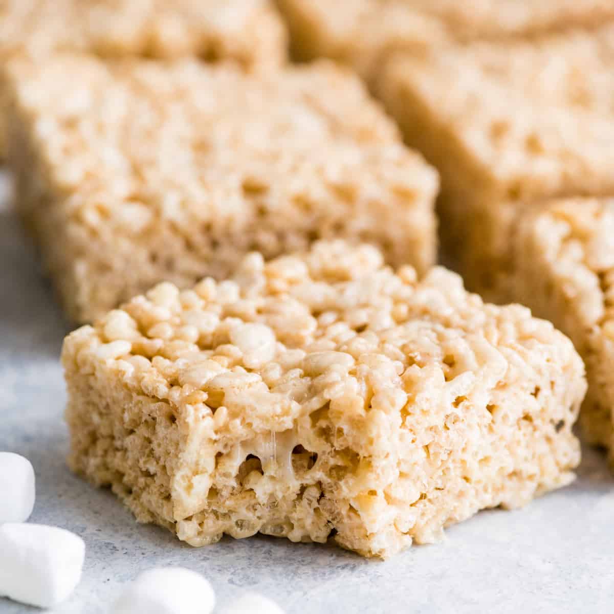 front view of a Rice Crispy Treat with a bite taken out of it, and 5 other rice crispy treats in the background and mini marshmallows in the foreground.