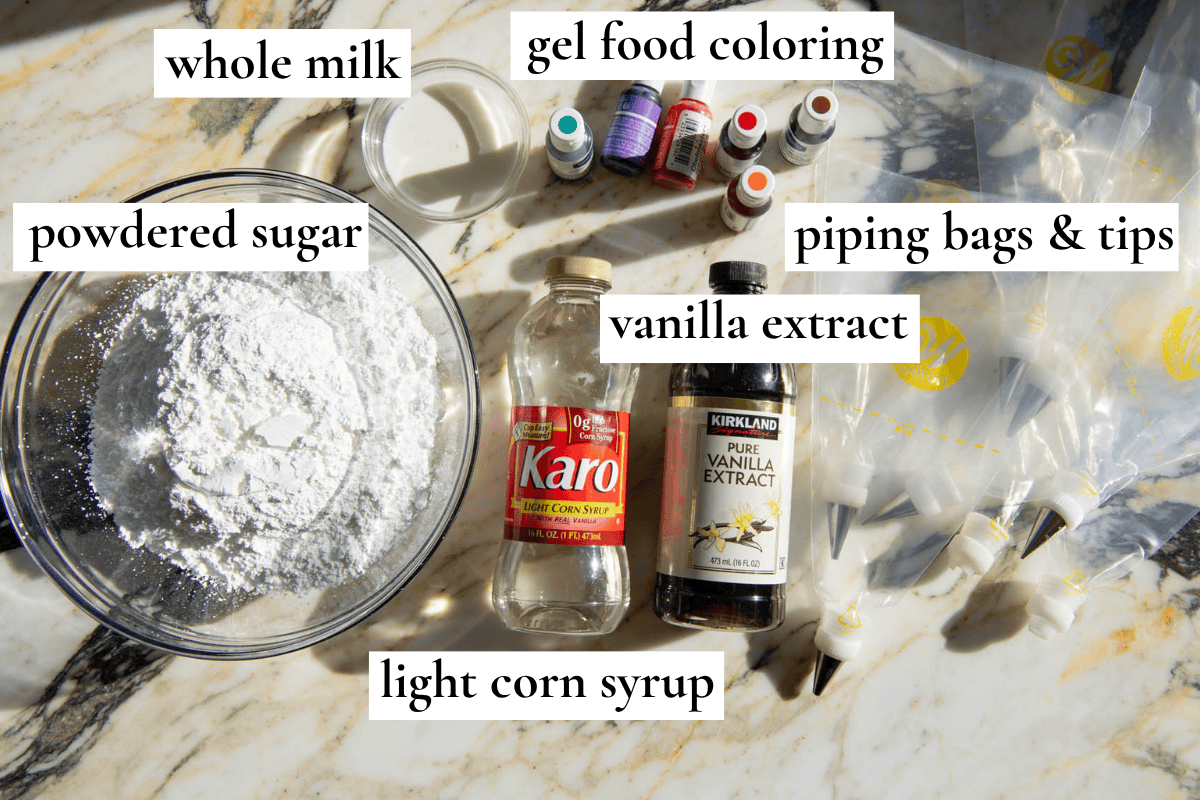overhead photo of the ingredients in this sugar cookies recipe cut out all labeled, the ingredients are powdered sugar in a glass bowl, whole milk in a small glass bowl, karo corn syrup in a bottle, a bottle of vanilla extra, bottles of gel food coloring and clear piping bags with tips.