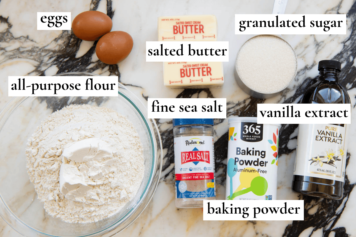 overhead photo of the ingredients in this sugar cookies recipe cut out all labeled, the ingredients are flour in a glass bowl, two brown eggs, two sticks of salted butter, granulated sugar in a silver measuring cup, a bottle of vanilla extract, baking powder and sea salt all on a marble surface