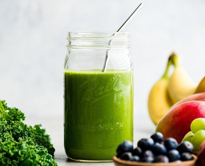 front view of a green smoothie in a large glass mason jar with a metal straw, surrounded with kale, blueberries, bananas, mangoes and green grapes