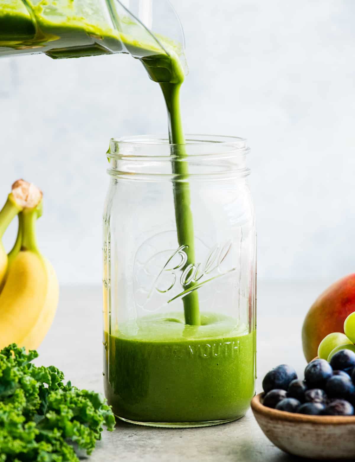 front view of a green smoothie being poured into a large glass mason jar, surrounded with kale, blueberries, bananas, mangoes and green grapes