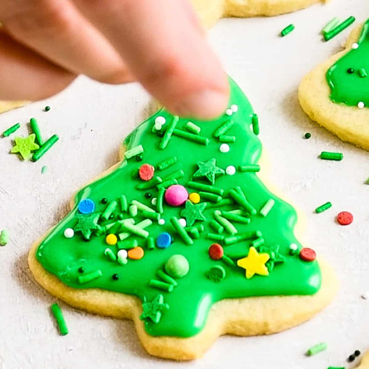 a hand putting sprinkles onto a green frosted Christmas tree cut out sugar cookie
