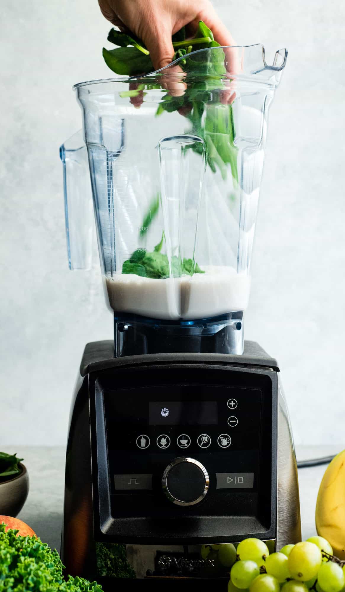 front view of a hand adding spinach to the almond milk in the container of a Vitamix A3500 blender to make this Green Smoothie Recipe