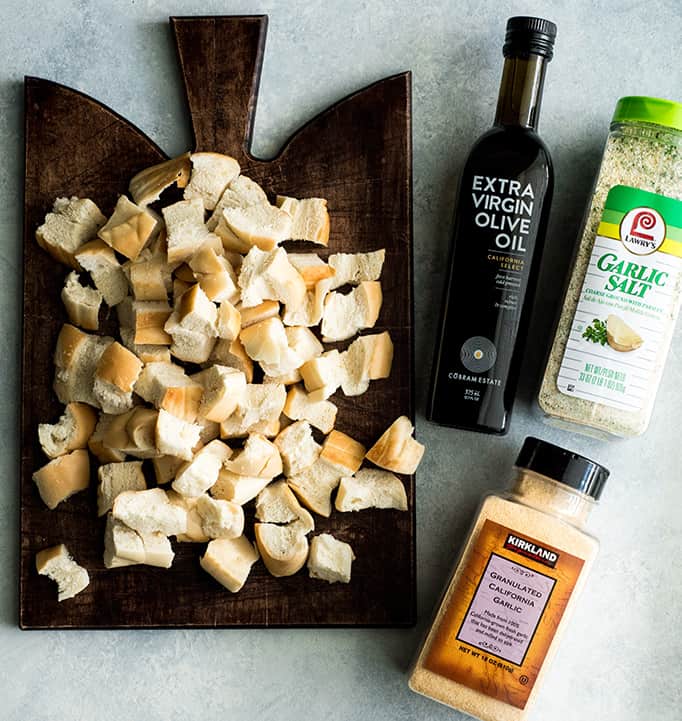 overhead view of the ingredients in this Homemade Crouton recipe: bread cubes on a square wood cutting board, a bottle of olive oil, a container of garlic salt and a container of garlic powder