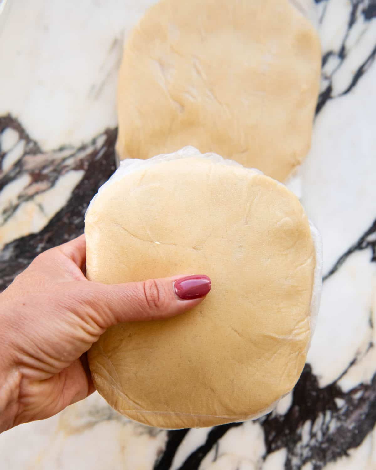 making cut out sugar cookies recipe - a hand holding a disc of sugar cookie dough wrapped in plastic wrap. There's a second disc of dough wrapped in plastic wrap on a marble surface below the first one.