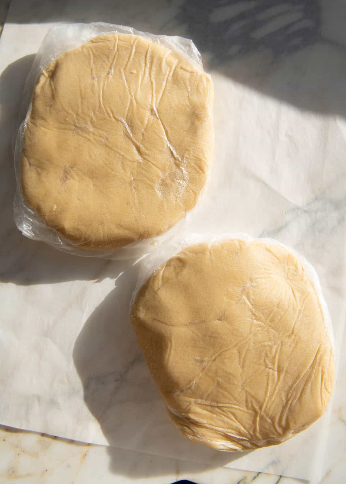 making cut out sugar cookies recipe - two chilled discs of dough wrapped in plastic wrap on parchment paper on a marble surface