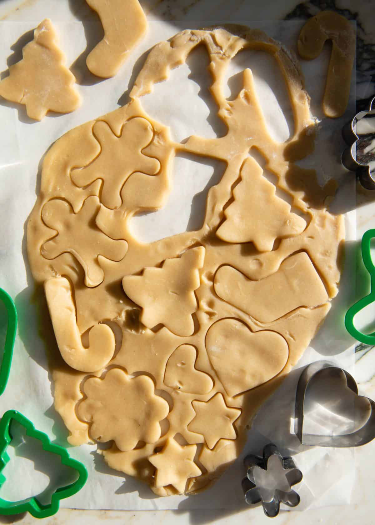 making cut out sugar cookies recipe - rolled sugar cookies dough on a marble surface with shapes cut out of it with cookie cutters like gingerbread men, Christmas trees, candy canes, hearts, stars and a flower.