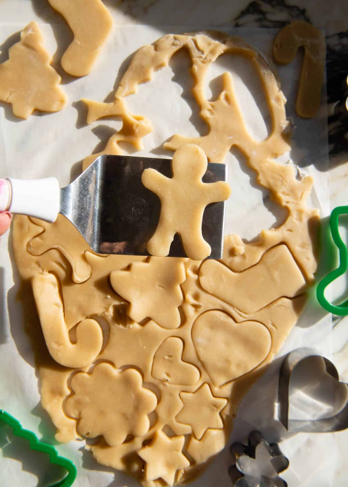 making cut out sugar cookies recipe - a metal spatula holding a gingerbread man shaped sugar cookie over the rest of the dough with shapes cut out of it