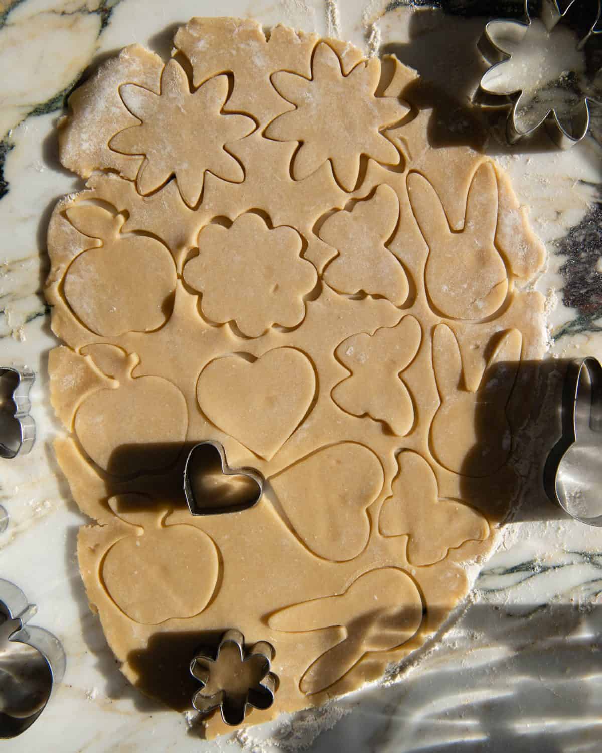 making cut out sugar cookies recipe - rolled sugar cookies dough on a marble surface with shapes cut out of it with cookie cutters like flowers, apples, butterflies, bunnies, and hearts.