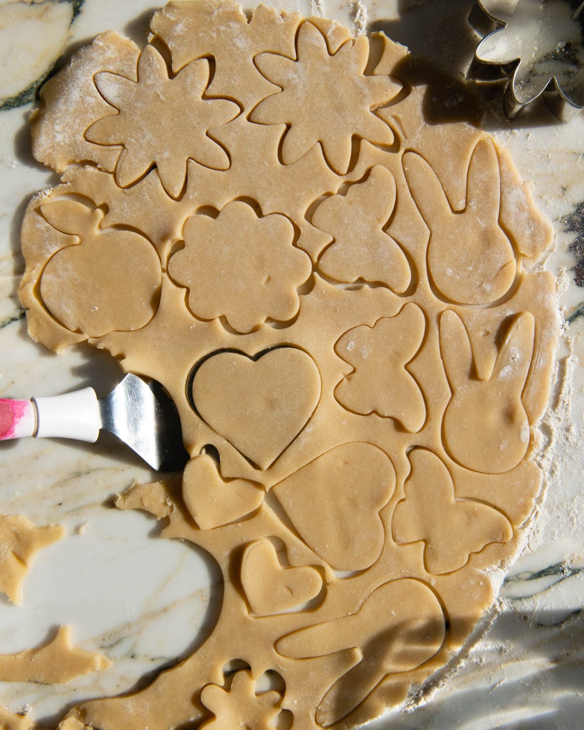 making cut out sugar cookies recipe - a metal spatula underneath a heart-shaped sugar cookie. there's many other shapes cut out of the rolled sugar cookie dough like bunnies, apples and flowers.