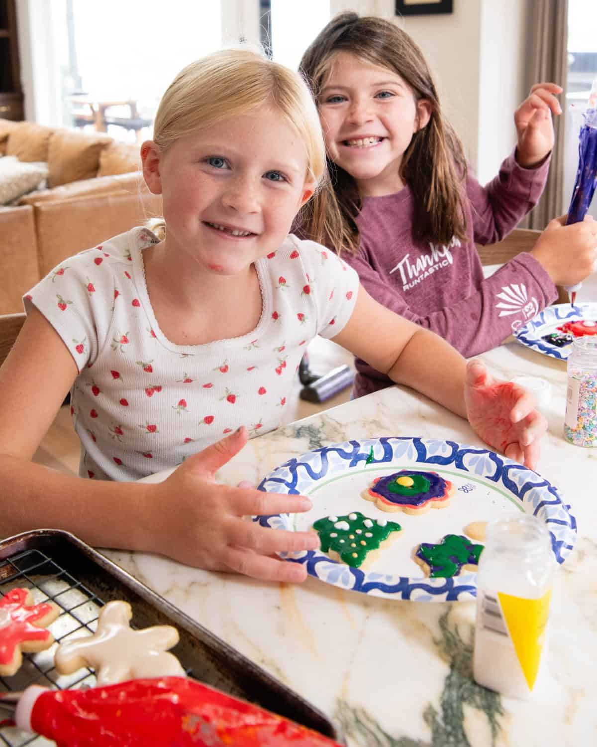two girls smiling while they frost cut out sugar cookies with sugar cookie icing