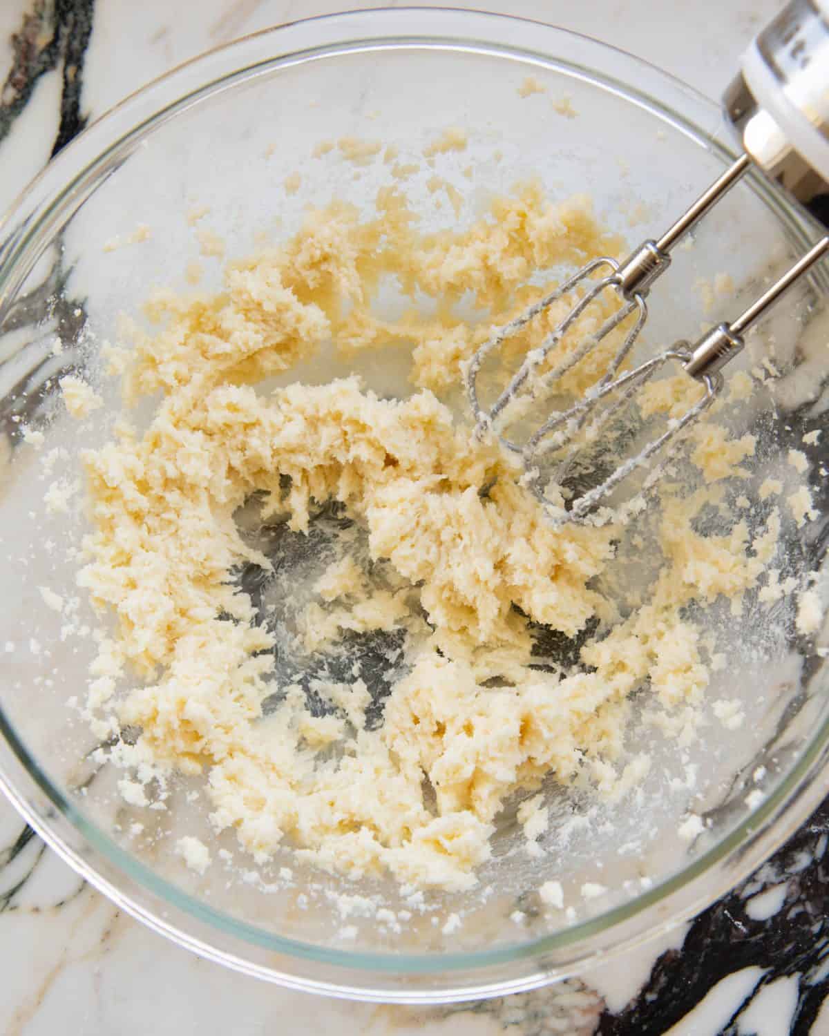 making cut out sugar cookies recipe - beaten butter and sugar in a large glass mixing bowl on a marble surface with a white handheld mixer with two beaters in the bowl