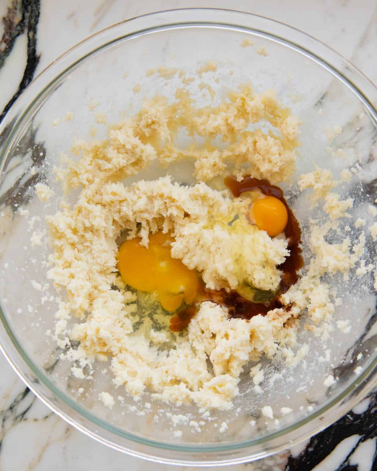making cut out sugar cookies recipe - two eggs and vanilla extract added to the beaten butter and sugar mixture in a large glass mixing bowl on a marble surface