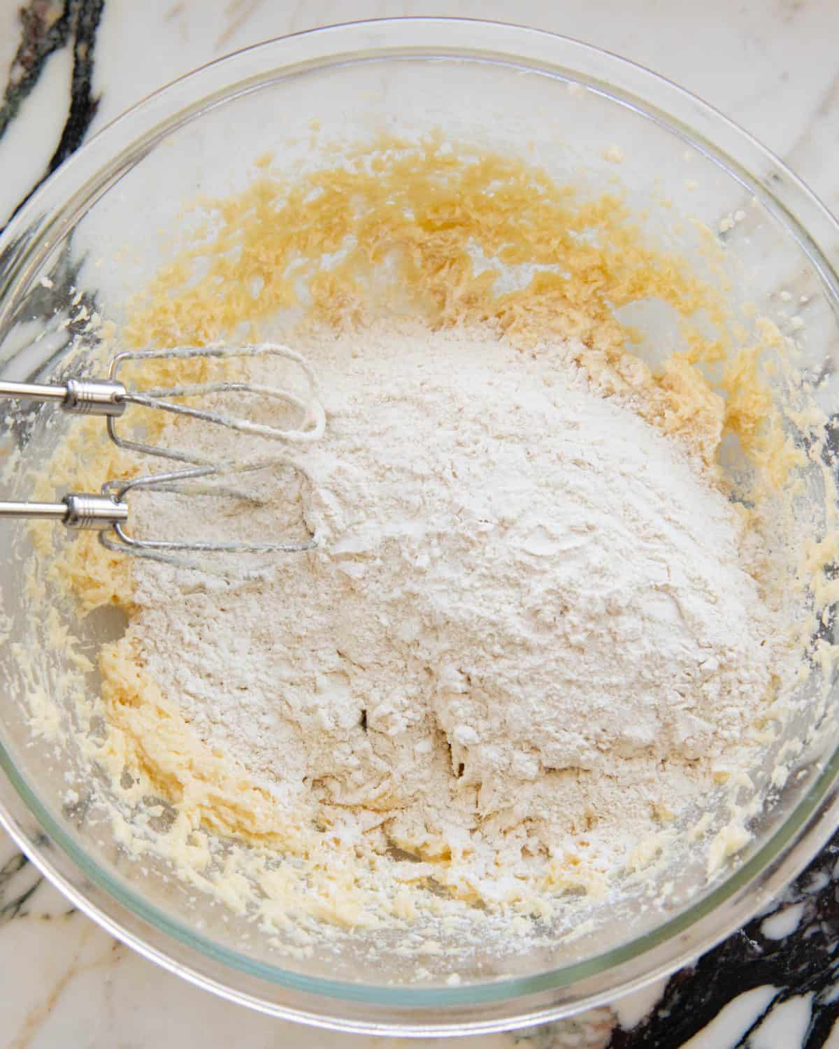 making cut out sugar cookies recipe - dry ingredients added to the beaten wet ingredients in a large glass mixing bowl on a marble surface, there are two beaters from a handheld mixer also in the bowl